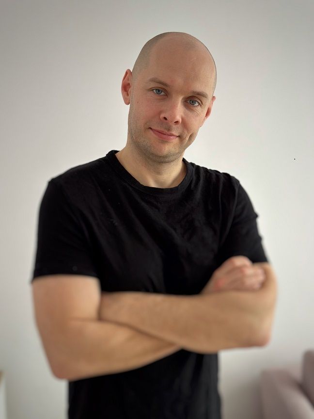 Marcin Moska — portret, ręce skrzyżowane, czarna koszulka, jasne tło